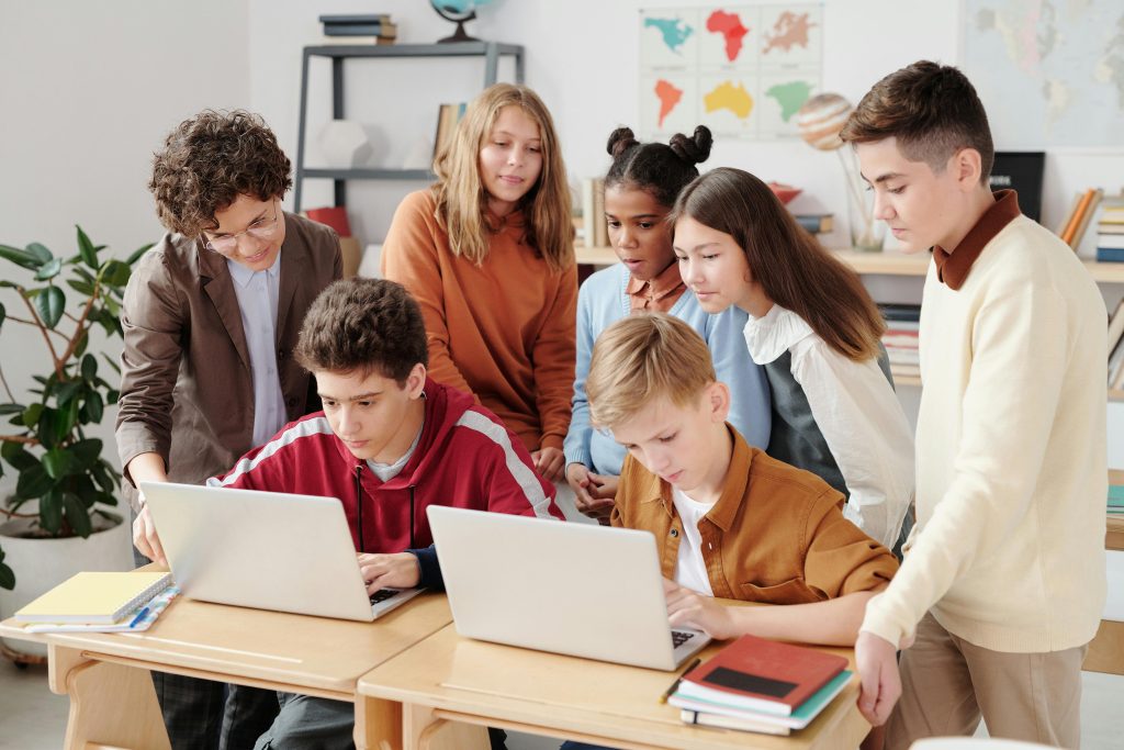 pexels-photo-5212687-5212687 A group of diverse students collaborating on laptops in a classroom setting, guided by a teacher.
